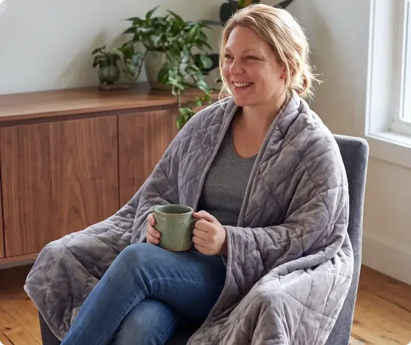 Woman relaxing in an armchair wrapped in the Velin Weighted Blanket with a mug of tea